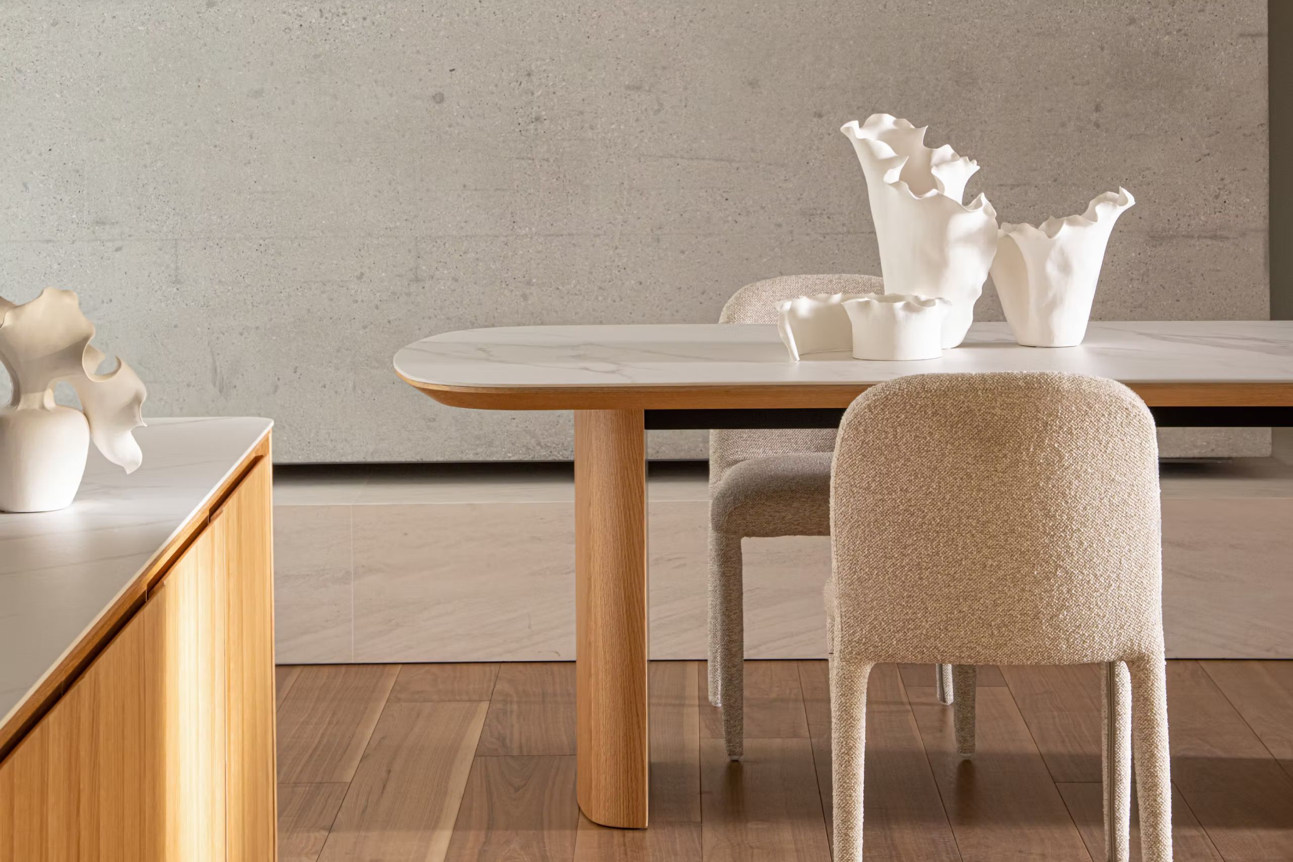 Mesa de madera con superficie clara, sillas tapizadas y jarrones blancos ante una pared mineral