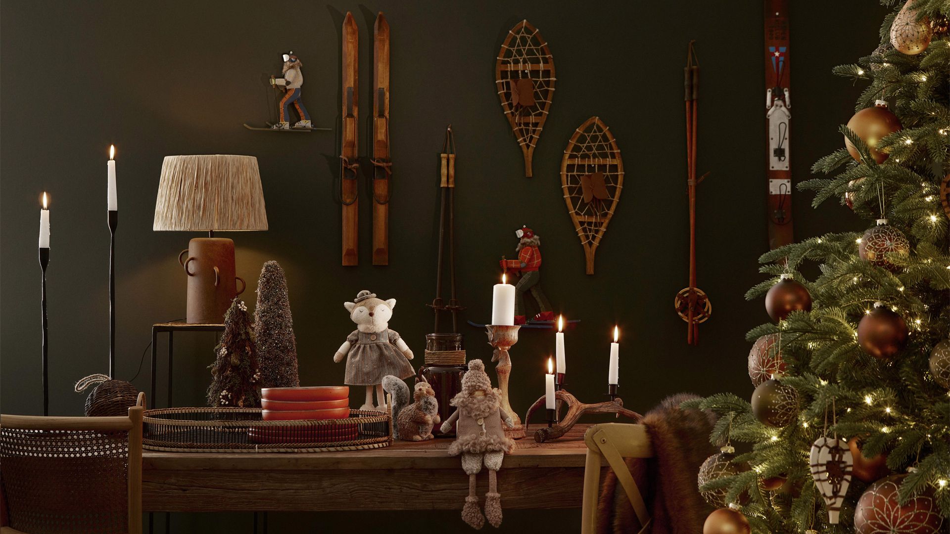 Mesa con velas encendidas, figuras navideñas y un árbol iluminado ante una pared verde oscuro