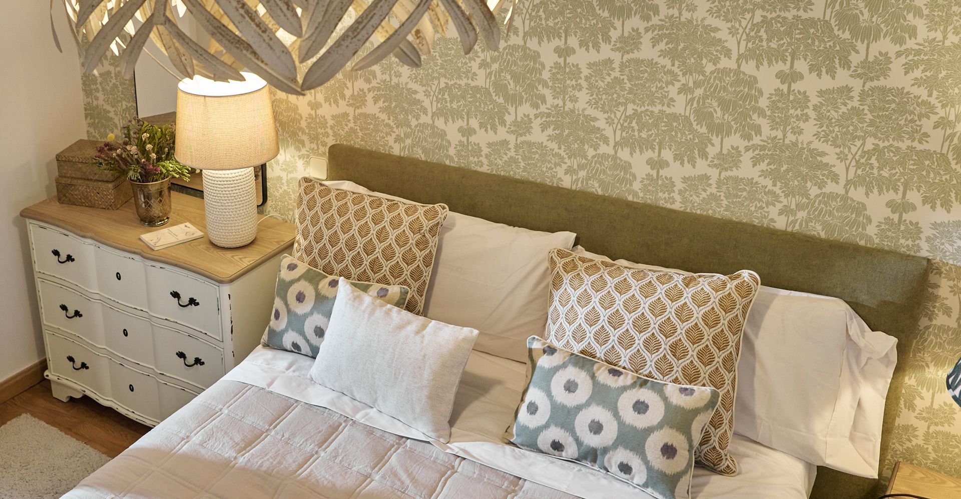 Cama con cabecero tapizado, cojines estampados y una cómoda blanca con lámpara ante un papel pintado de árboles